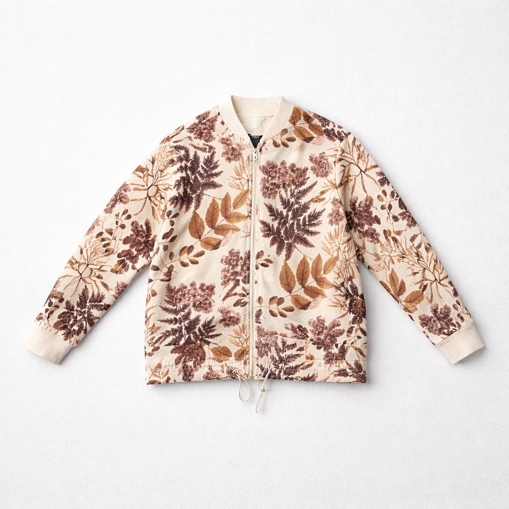 Chaqueta de color crema con cremallera en la parte delantera y cordón de ajuste en el bajo, con estampado botánico de hojas marrones y tostadas sobre fondo blanco.