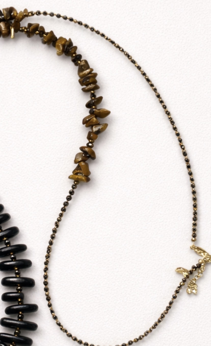 El Collar largo de Ébano y Ojo de Tigre (90-96 cm) luce virutas irregulares marrones, cadena dorada con cuentas finas y pequeño dije dorado, exhibido sobre una superficie clara y texturizada.