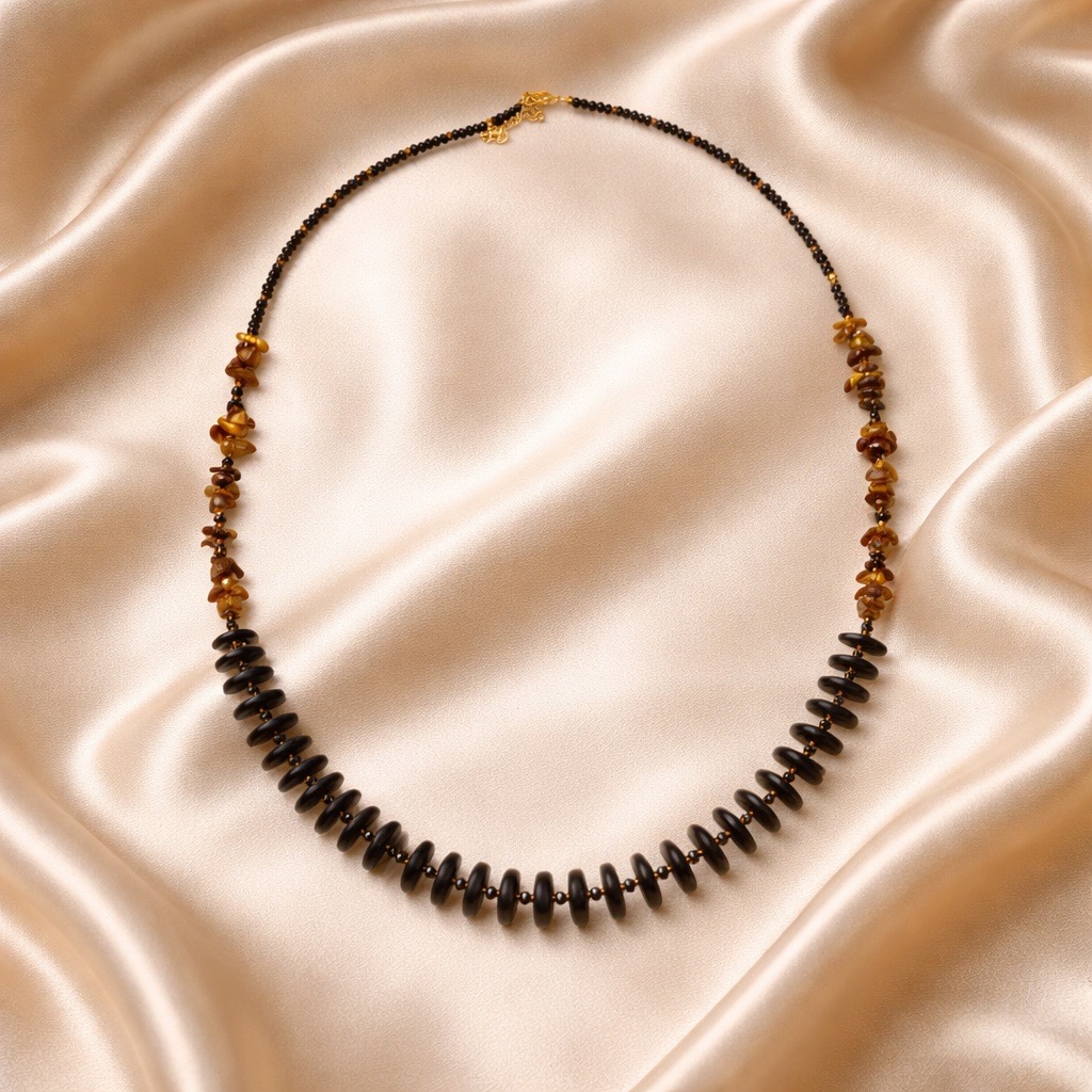 El cuello largo de Ébano y Ojo de Tigre (90-96 cm) con cuentas alternas en negro y ámbar se exhibe sobre un fondo suave de satén color champán.