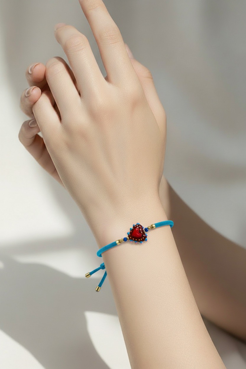 Una mano luce la Pulsera Corazón de cristal con cordón azul, que exhibe un charm de cristal rojo en forma de corazón con detalles dorados sobre un fondo suave y neutro.