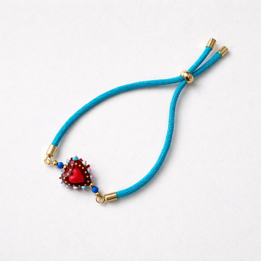 La Pulsera Corazón de cristal con cordón azul tiene un cordón azul dorado y un colgante rojo en forma de corazón adornado con cuentas de colores, sobre un fondo blanco.