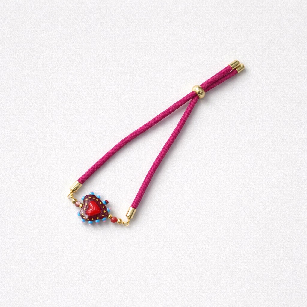 La Pulsera Corazón de cristal con cordón azul es una pulsera de cordón azul con un charm de corazón de cristal y detalles de abalorios, acabada con cierres dorados, que se muestra sobre un fondo blanco.