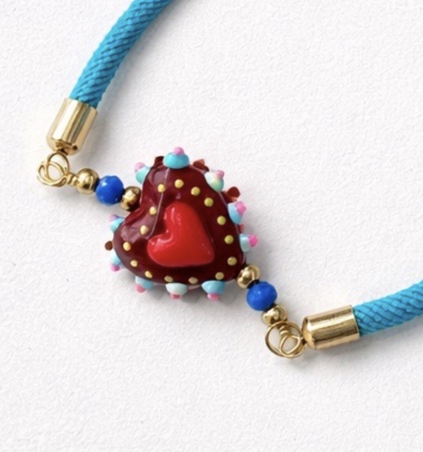 Un primer plano de la Pulsera Corazón de cristal con cordón azul: una pulsera de cordón azul con cierres dorados, con una cuenta central de cristal en forma de corazón en rojo y marrón, rodeada de puntos de colores y dos cuentas redondas azules a cada lado.
