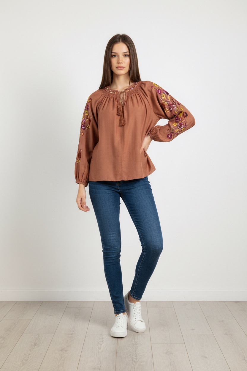 Una mujer, de pie contra una pared blanca y lisa, lleva una blusa marrón con motivos florales bordados en las mangas -una elección perfecta para quienes buscan blusas y camisas para mujer- combinada con vaqueros pitillo azules y zapatillas blancas.