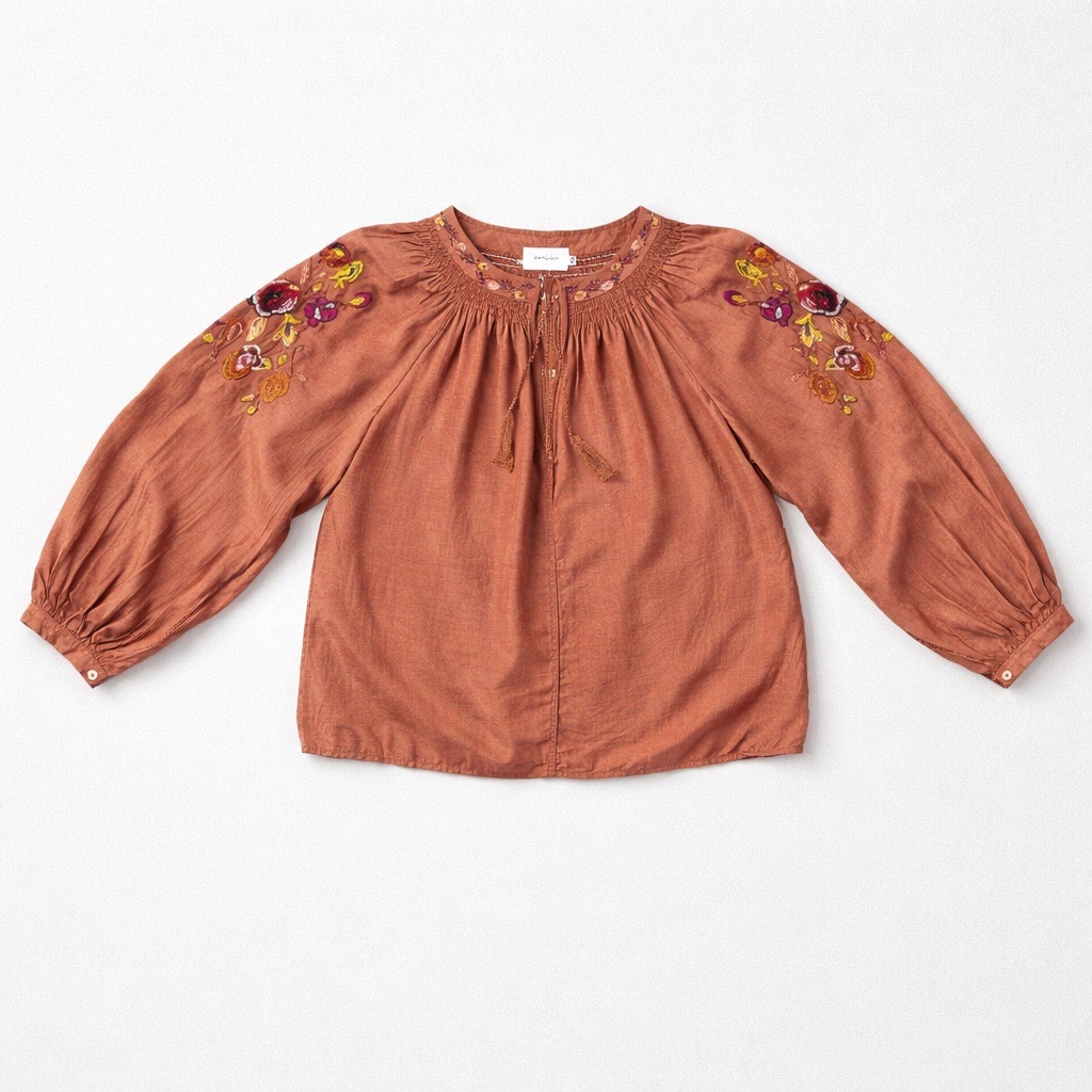 Blusa de manga larga color óxido con motivos florales bordados en los hombros, cuello fruncido con lazos y botones en los puños, colocada en plano sobre fondo blanco.