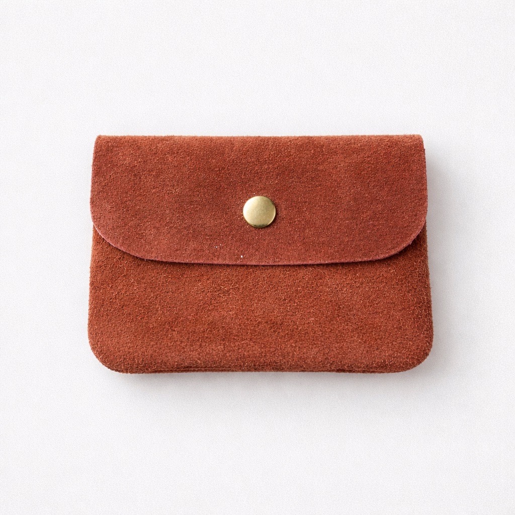 Sobre un fondo blanco liso se coloca una pequeña bolsa rectangular de ante marrón con solapa redondeada, sujeta por un único botón dorado.