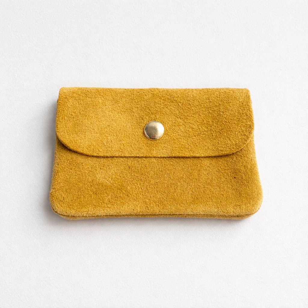 Una pequeña bolsa de ante amarillo mostaza con cierre de solapa y un único botón a presión metálico, colocada sobre un fondo blanco liso.