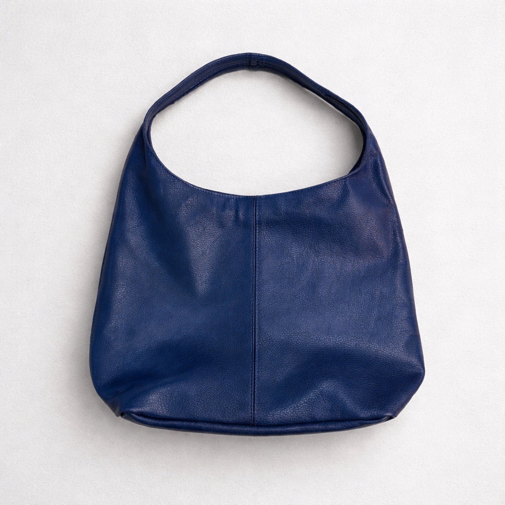 Bolso de piel azul oscuro con una sola correa ancha para el hombro, sobre un fondo claro y neutro. El bolso tiene un diseño sencillo, sin adornos, y una forma ligeramente redondeada.