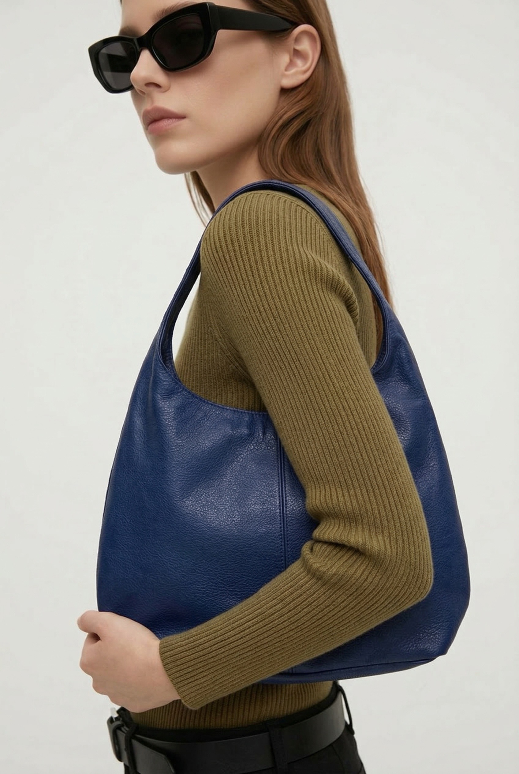 Una mujer con gafas de sol negras y jersey de canalé verde oliva sostiene un gran bolso de piel azul oscuro. Está de pie sobre un fondo liso y claro y mira de reojo.