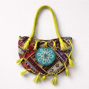Colorido bolso bohemio con estampado de patchwork, asas trenzadas amarillas, borlas amarillas de piel y un gran diseño bordado turquesa en el centro, sobre fondo blanco.